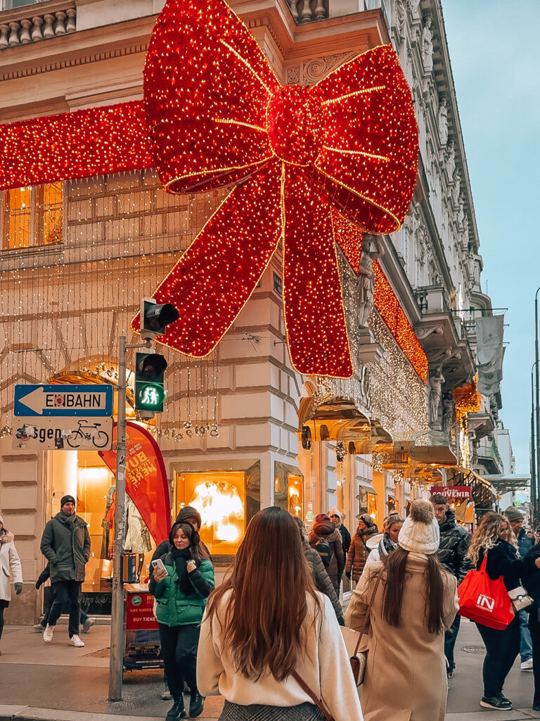 Close-up street-level view of Vienna’s iconic oversized red Christmas bow lit up with twinkling lights, as holiday shoppers walk beneath festive decorations and shopfronts.

