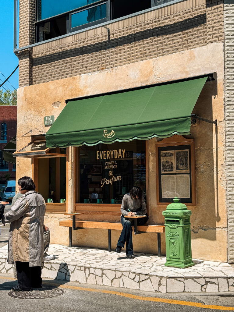 Charming café styled like an old post office with a green awning and vintage mailbox, where a woman writes in her notebook on a bench.
