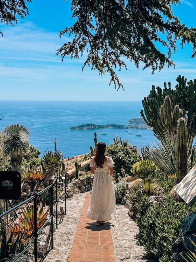 A woman in a white dress walks down a stone path lined with cacti and succulents in the Exotic Garden of Èze, overlooking the sparkling Mediterranean Sea.
