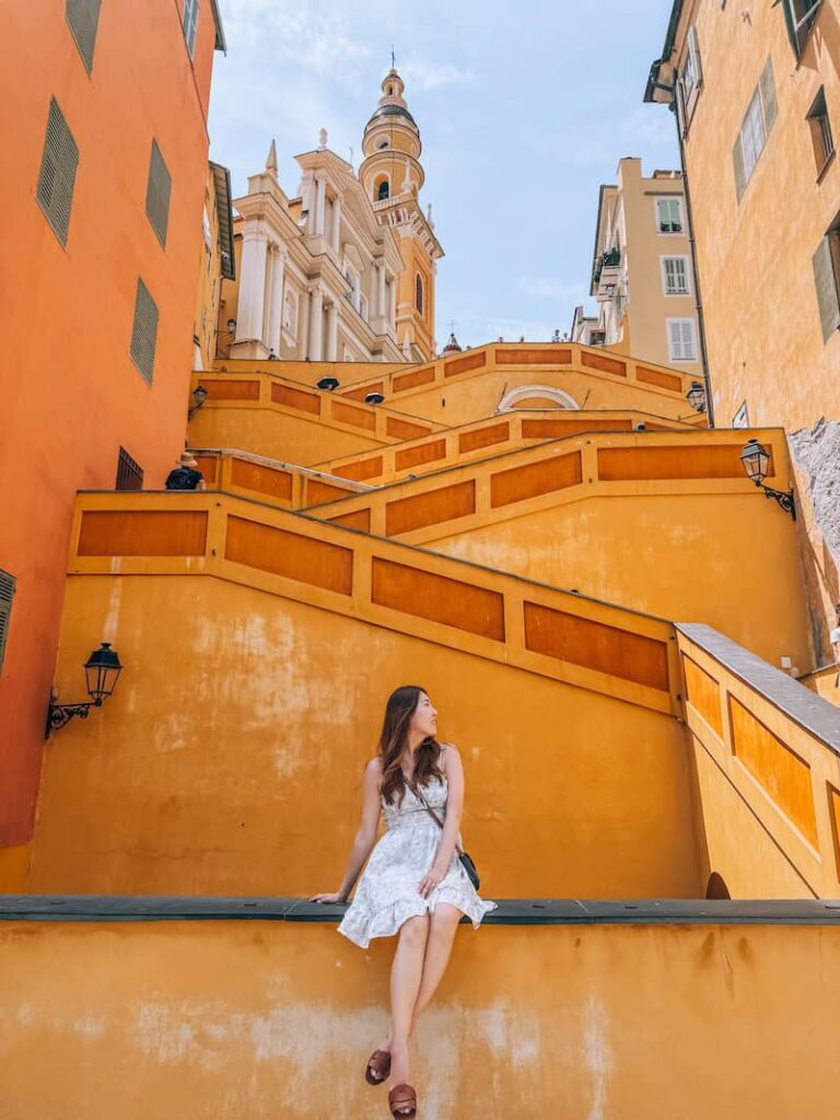A woman in a white dress sits on a bright yellow wall in front of a zigzag staircase and pastel buildings, with the Baroque-style bell tower of Menton’s Basilica peeking above.
