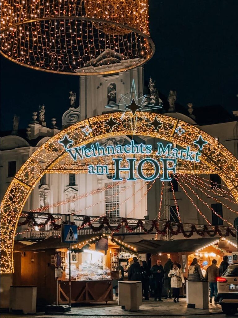 Nighttime view of the illuminated entrance arch at the Am Hof Christmas Market in Vienna, decorated with glowing stars and festive string lights above the surrounding stalls.