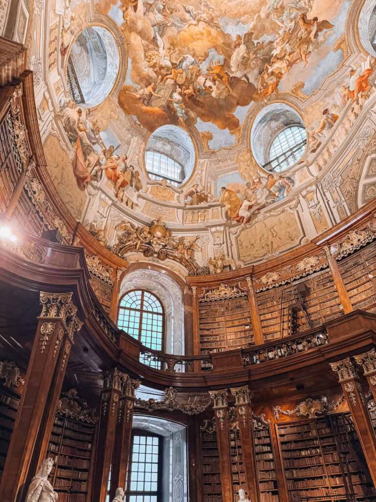 Baroque-style library interior with towering wooden bookshelves, ornate gold detailing, and a stunning frescoed ceiling depicting dramatic scenes of angels and clouds, located in the Austrian National Library in Vienna.