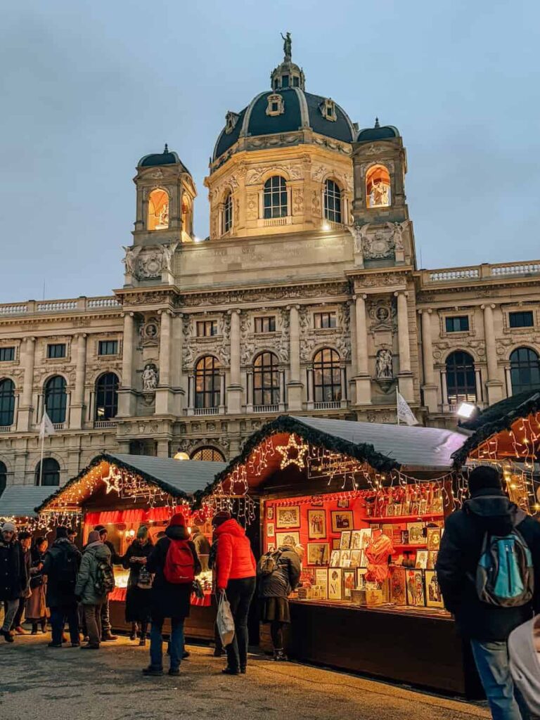 People browsing Christmas market stalls decorated with fairy lights in front of the illuminated Natural History Museum at Maria-Theresien-Platz in Vienna.