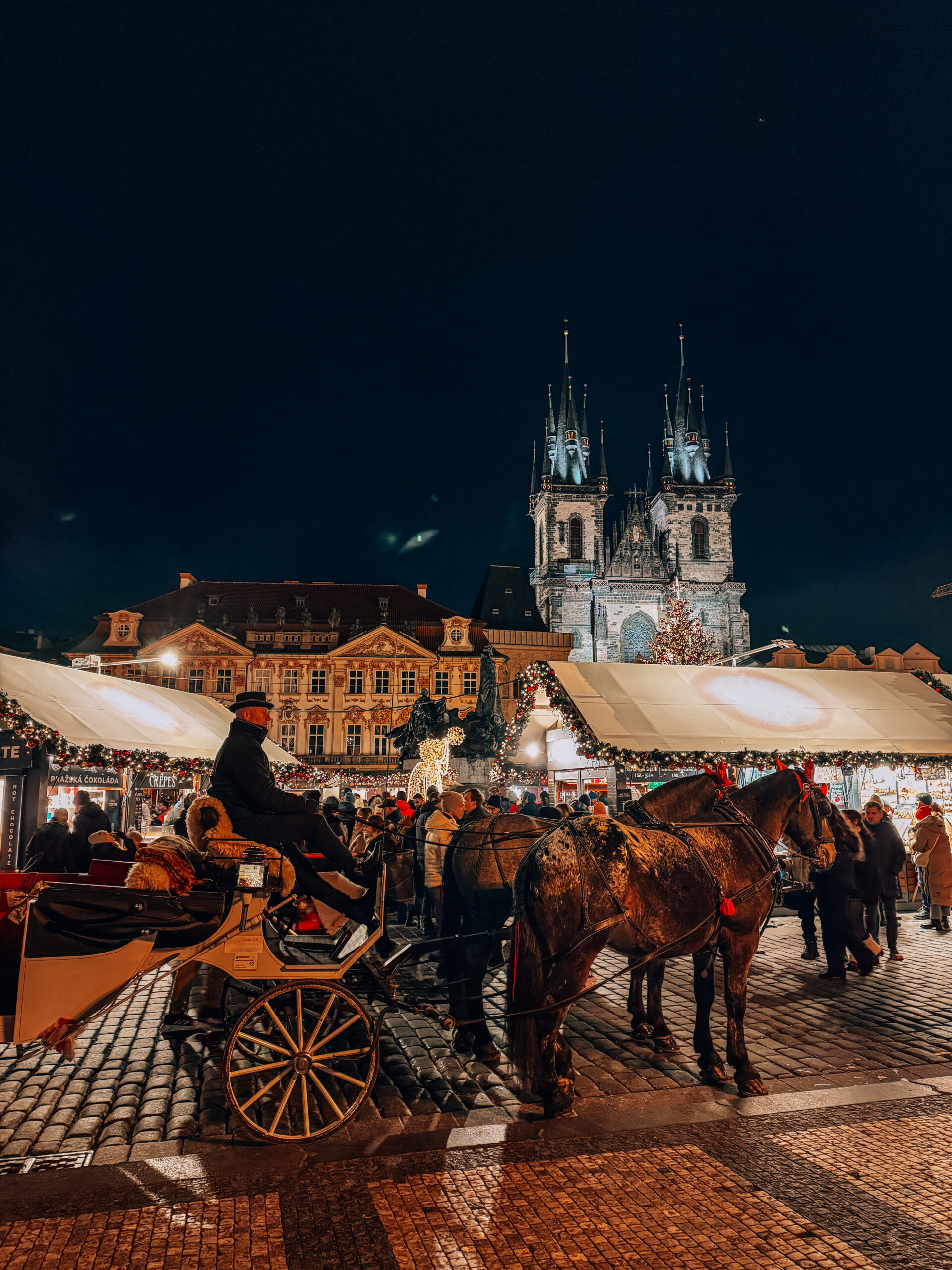 Horse-drawn carriage in front of festive Christmas market stalls in Prague’s Old Town Square at night, with the Church of Our Lady before Týn illuminated in the background and crowds enjoying the holiday atmosphere during Christmas markets in Prague