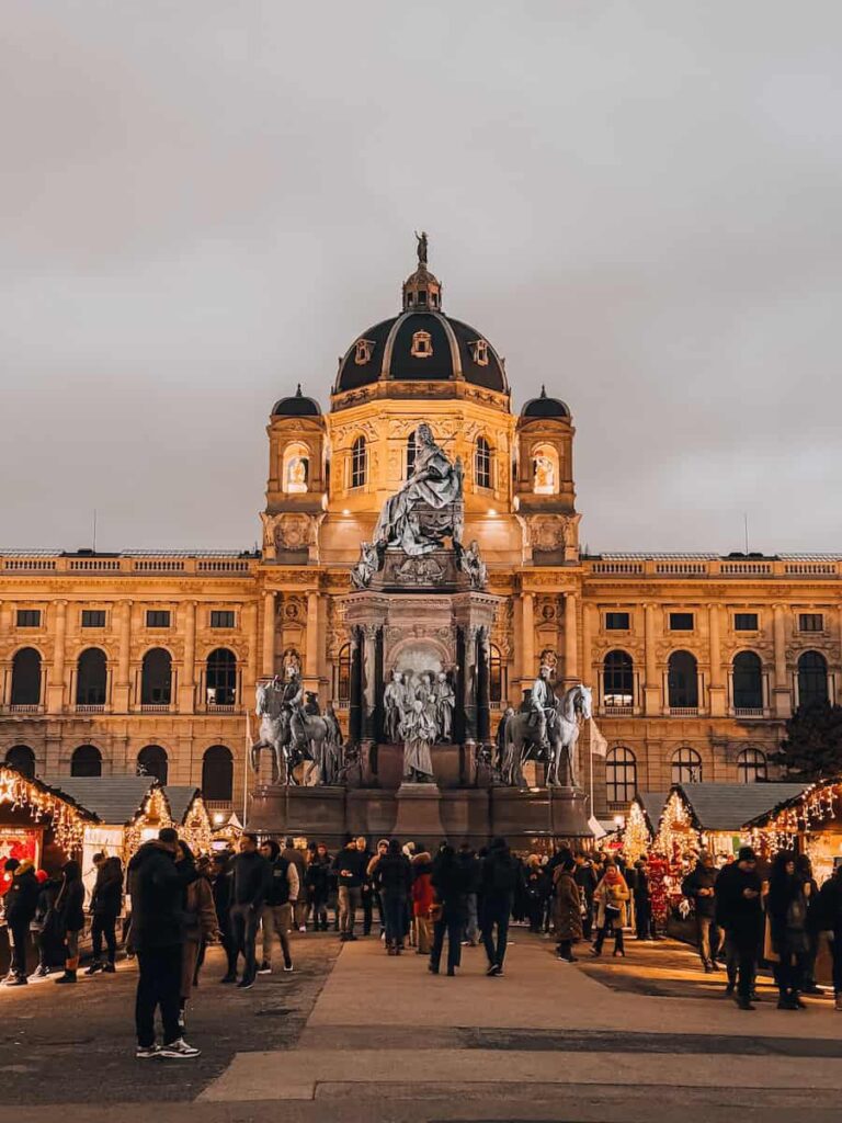 Crowds gather at the Christmas market in Vienna’s Maria-Theresien-Platz, with twinkling lights, festive stalls, and the illuminated Natural History Museum in the background.