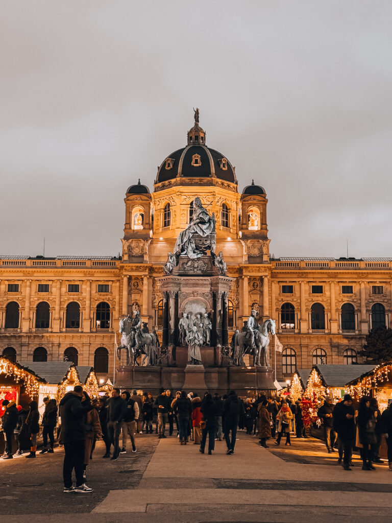 Crowds gather at the Christmas market in Vienna’s Maria-Theresien-Platz, with twinkling lights, festive stalls, and the illuminated Natural History Museum in the background.