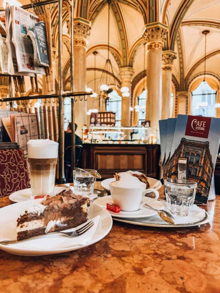 A cozy table setting at Café Central in Vienna featuring a cappuccino with whipped cream, a latte, glasses of water, a slice of layered chocolate cake, and elegant brochures. The background reveals ornate vaulted ceilings, arched windows, and warm lighting inside the historic Viennese coffeehouse.