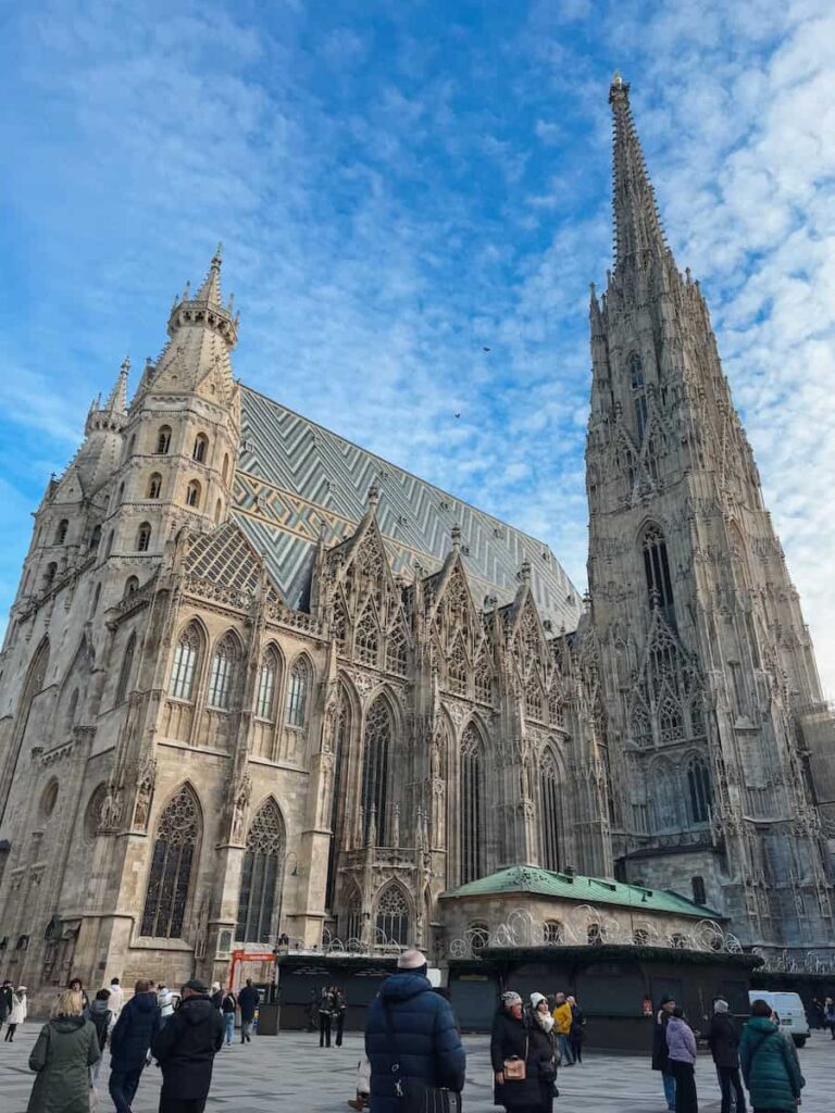 Exterior view of St. Stephen’s Cathedral (Stephansdom) in Vienna, showcasing its intricate Gothic architecture, tall South Tower, and colorful patterned tiled roof. Visitors gather in the plaza beneath the cathedral on a clear winter day.