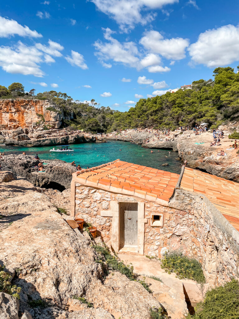 A small stone boathouse with a terracotta roof sits on rocky cliffs overlooking the turquoise waters of Cala S'Almunia in Mallorca, Spain. Sunbathers and swimmers enjoy the scenic cove surrounded by pine trees and rugged cliffs under a bright blue sky with scattered clouds.