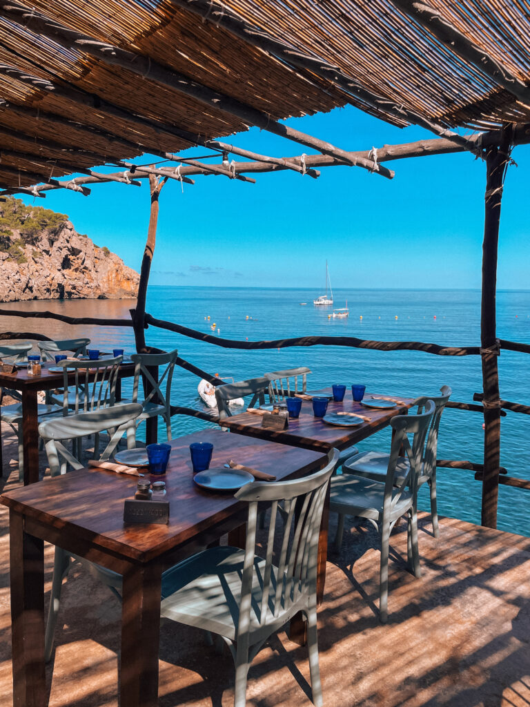 Outdoor dining tables with blue glasses and white plates at a rustic seaside restaurant in Cala Deià, Mallorca, overlooking the deep blue Mediterranean Sea and anchored sailboats.