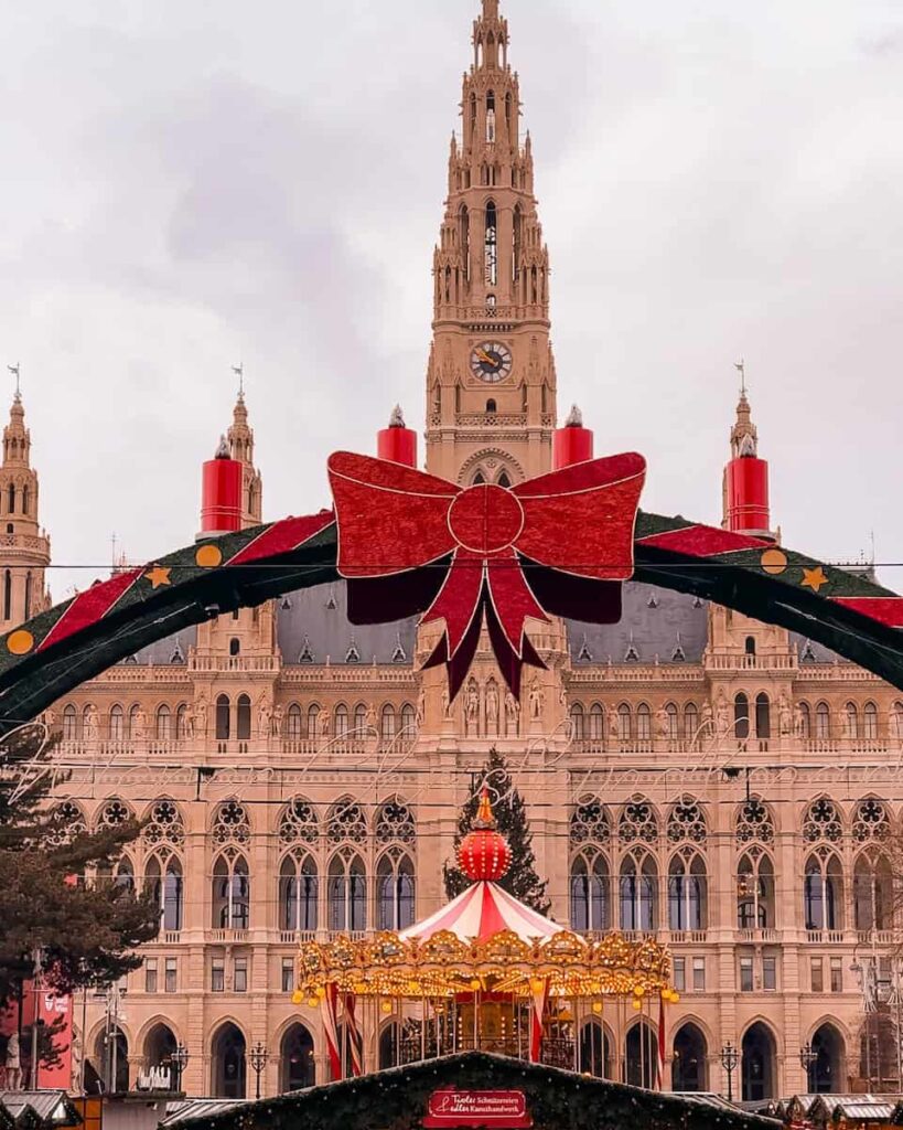 Christmas market at Vienna’s Rathausplatz featuring a bright red arch with a bow and candles, a festive carousel with golden lights, and the grand Vienna City Hall building in the background under an overcast winter sky.