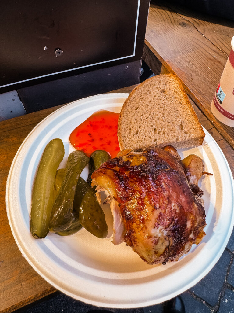 A hearty meal of roasted chicken, sliced pickles, rye bread, and red sauce served on a paper plate at a Prague Christmas market stall.