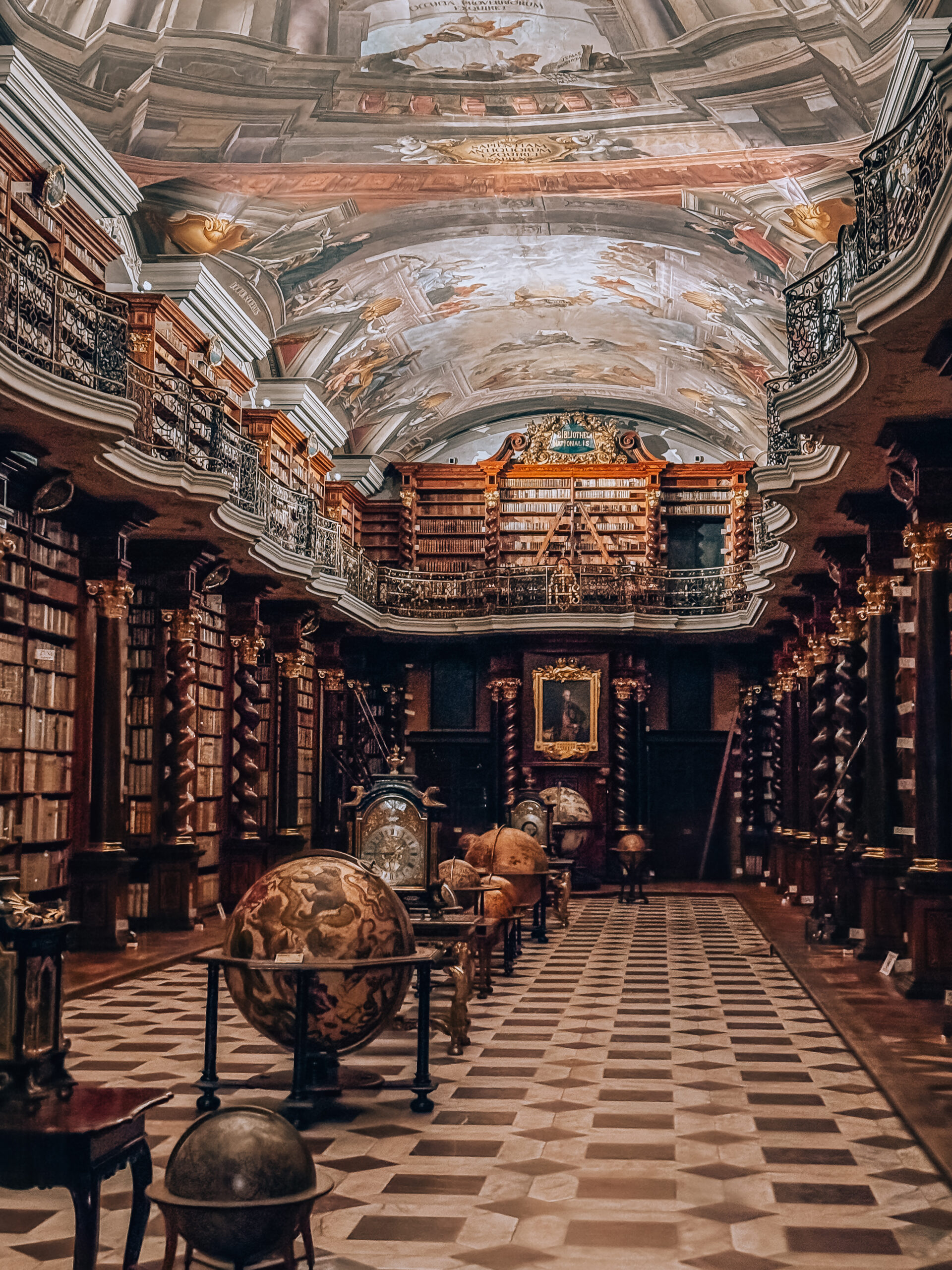 The Baroque Library Hall at the Klementinum in Prague, featuring ornate ceiling frescoes, antique globes, astronomical instruments, and walls lined with historic books