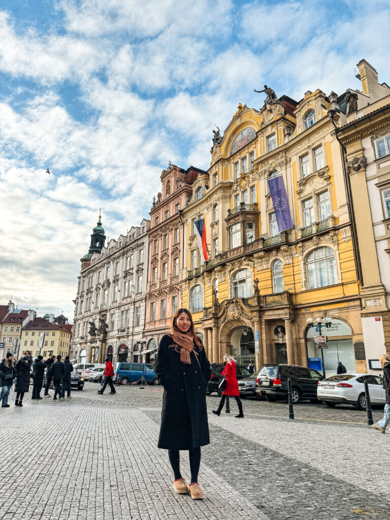 Woman standing in Prague’s Old Town Square with colorful Baroque and Rococo facades in the background on a bright winter day.