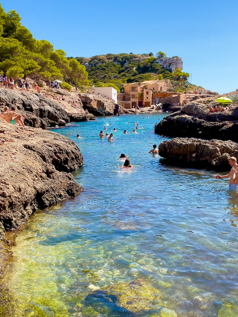 Swimmers enjoying the turquoise waters of Cala s'Almunia in Mallorca, framed by rocky cliffs and stone houses in the background, with clear shallows revealing rocks beneath the surface.