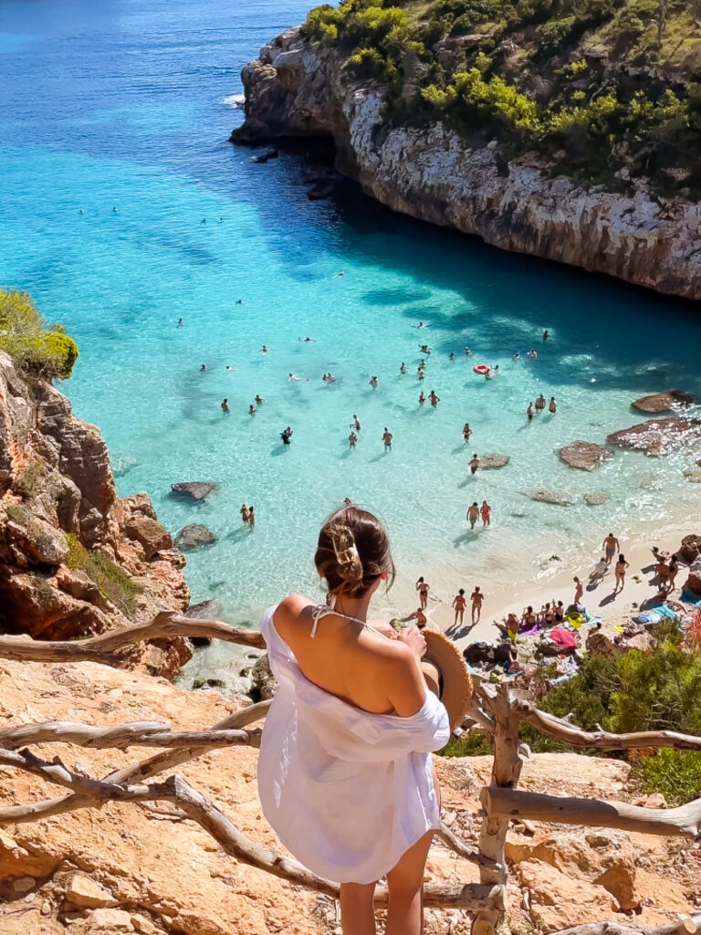 A woman wearing a wide-brimmed straw hat and a white cover-up stands at a cliffside viewpoint overlooking the turquoise waters and sandy beach of Cala des Moro in Mallorca, Spain, with swimmers scattered in the shallow water below.