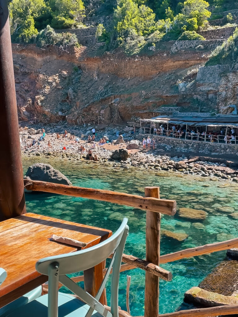 Table at a seaside restaurant in Cala Deià, Mallorca, with a view of the rocky beach below, where people are swimming and sunbathing beside crystal-clear turquoise water.