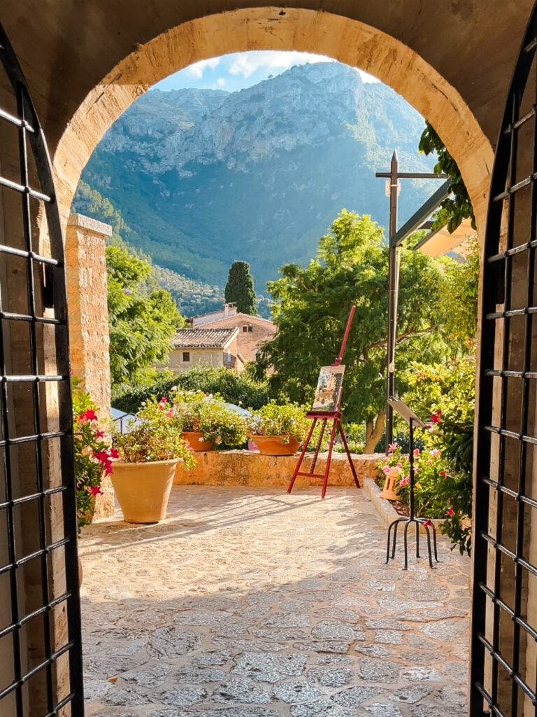 View through a stone archway framed by wrought-iron gates, looking out to a sunlit terrace with flower pots, an artist’s easel, and mountains rising in the background in Deià, Mallorca.
