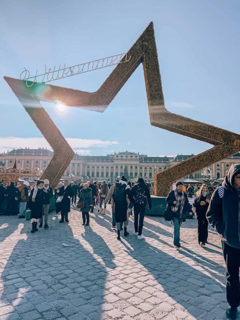 Visitors walking through the giant star-shaped “Willkommen” entrance at the Schönbrunn Palace Christmas Market on a sunny winter day in Vienna.