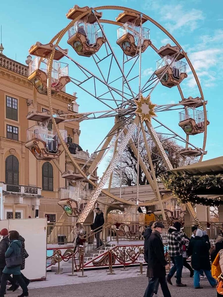 Vintage-style Ferris wheel at the Schönbrunn Christmas Market in Vienna, with people strolling past the festive palace courtyard beneath a bright blue winter sky.