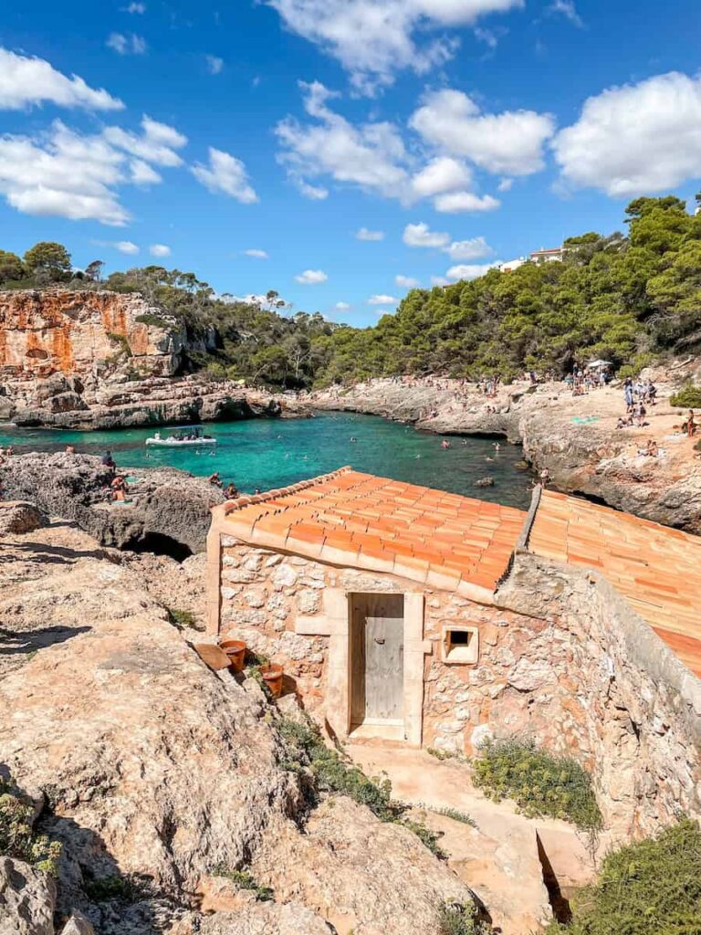 A small stone boathouse with a terracotta roof sits on rocky cliffs overlooking the turquoise waters of Cala S'Almunia in Mallorca, Spain. Sunbathers and swimmers enjoy the scenic cove surrounded by pine trees and rugged cliffs under a bright blue sky with scattered clouds.