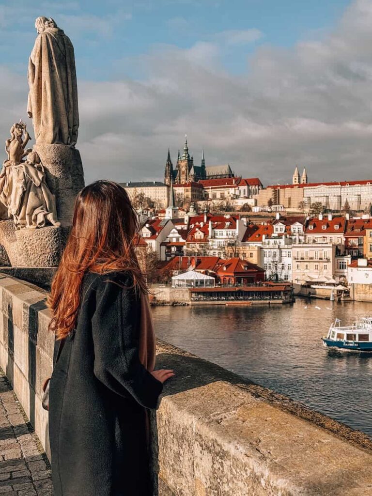 Woman in a long black coat standing on Charles Bridge in Prague, gazing across the Vltava River toward Prague Castle and red-roofed buildings under a partly cloudy sky