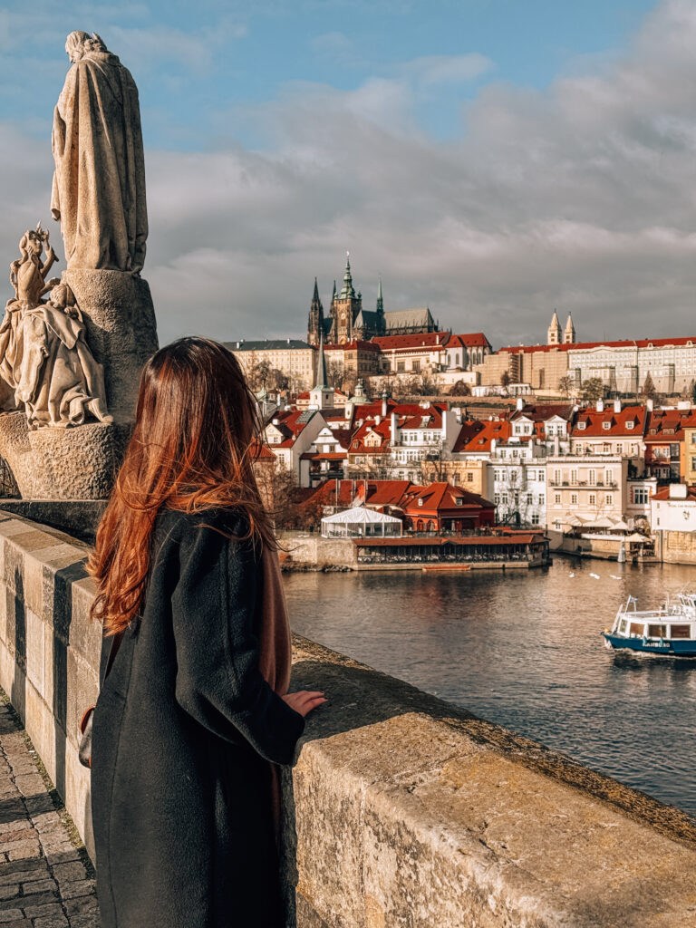 Woman in a long black coat standing on Charles Bridge in Prague, gazing across the Vltava River toward Prague Castle and red-roofed buildings under a partly cloudy sky