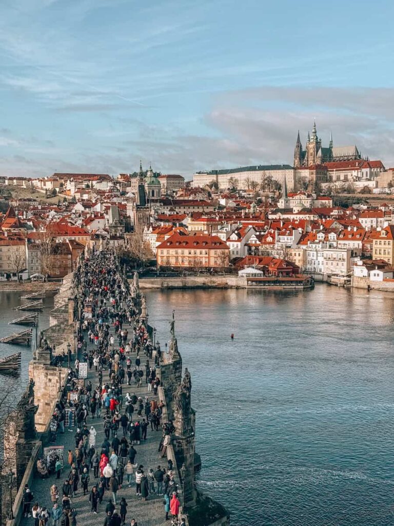 Bird’s-eye view of Charles Bridge in Prague filled with pedestrians, crossing the Vltava River with red-roofed buildings and Prague Castle in the background on a clear winter day