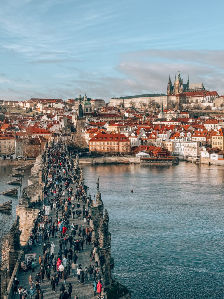 Bird’s-eye view of Charles Bridge in Prague filled with pedestrians, crossing the Vltava River with red-roofed buildings and Prague Castle in the background on a clear winter day
