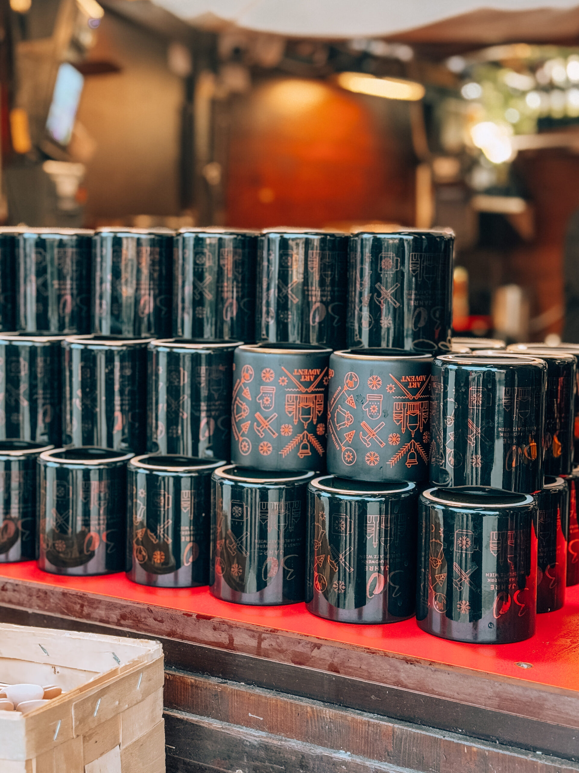 Stacked rows of matte black souvenir mugs with minimalist red designs at Art Advent Christmas Market in Karlsplatz, Vienna—displayed on a festive red counter.