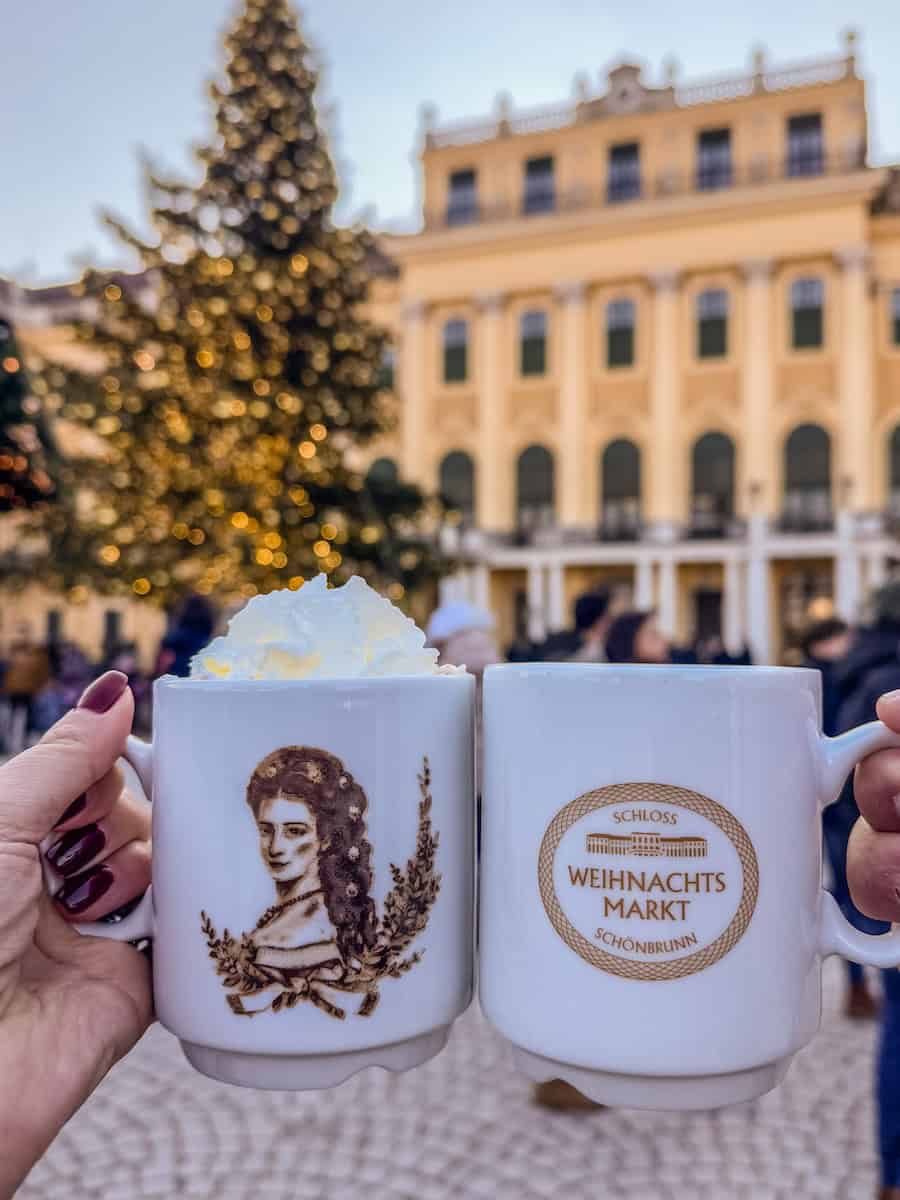 Close-up of two white mugs from the Schönbrunn Palace Christmas Marke. One featuring Empress Sisi and the other with the market’s logo, held up in front of the decorated palace and Christmas tree.