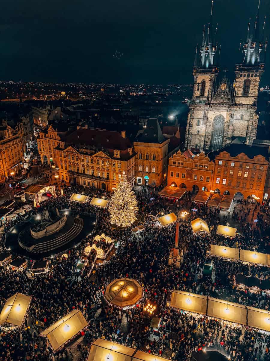 Aerial night view of Prague’s Old Town Square glowing with Christmas lights, showing the large Christmas tree in the center, surrounded by festive market stalls and crowds, with the twin towers of the Church of Our Lady before Týn rising above the square.