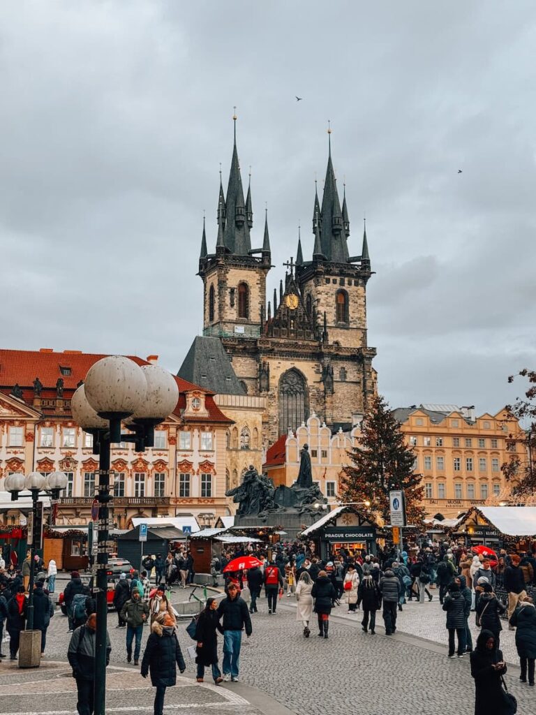 Daytime view of Prague’s Old Town Square during the Christmas market, with people walking among decorated wooden stalls and a Christmas tree in front of the Gothic Church of Our Lady before Týn under a cloudy sky.