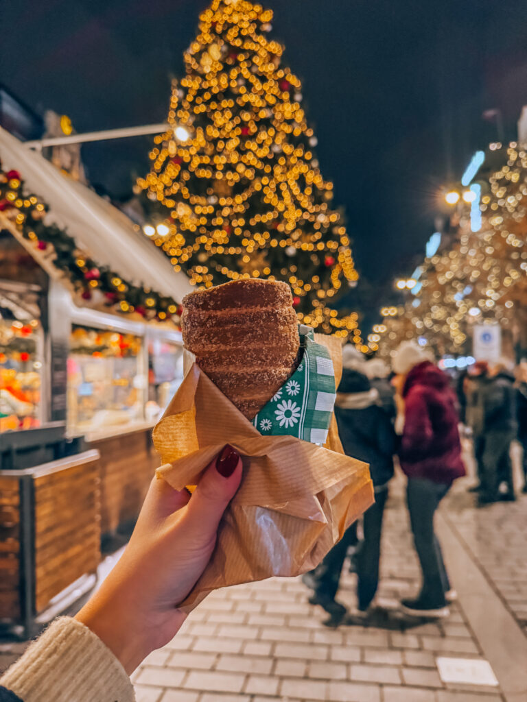 Close-up of a hand holding a freshly baked trdelník pastry at a Christmas market in Prague, with a large Christmas tree and blurred holiday lights in the background