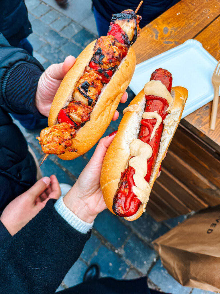 Two hands holding street food: one with a grilled chicken skewer in a bun and the other with a sausage hot dog topped with ketchup and mustard, set at a Prague Christmas market.