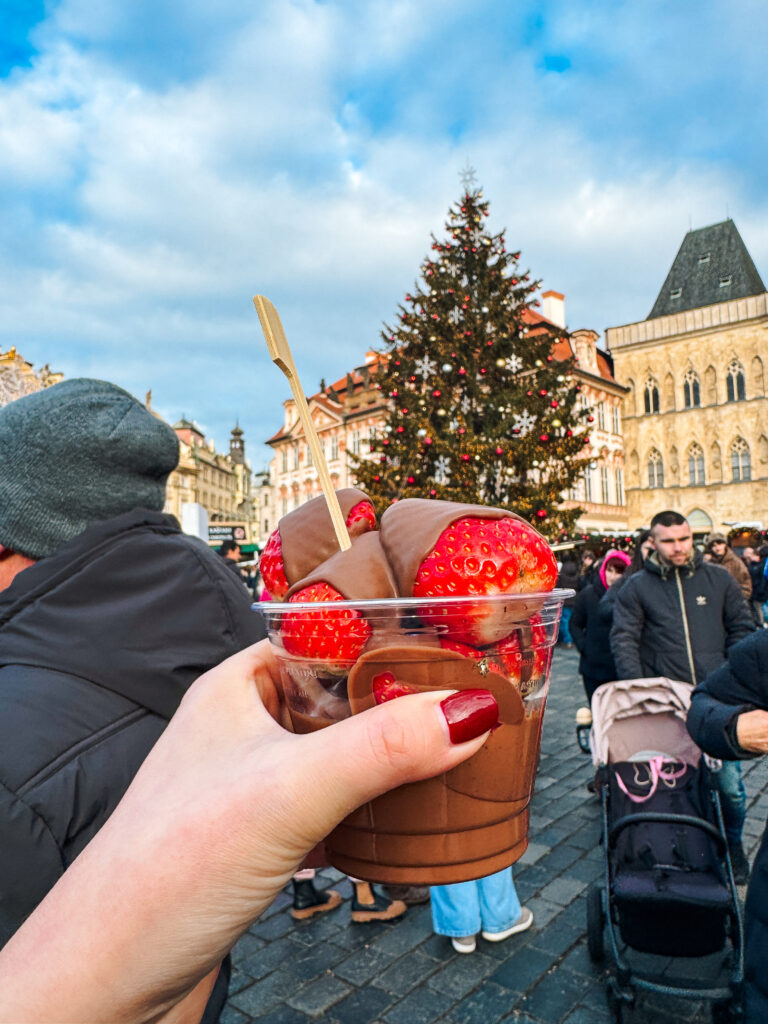 A cup of chocolate-dipped strawberries held up in front of a large decorated Christmas tree in Prague’s Old Town Square.