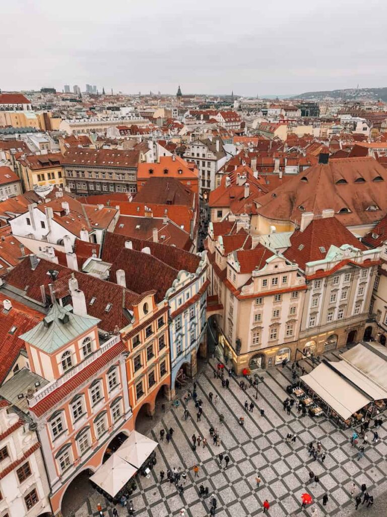 Aerial view of colorful historic buildings with red rooftops in Prague’s Old Town, with a cobblestone square below filled with people and café umbrellas on a cloudy winter day