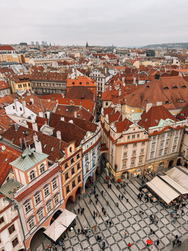 Aerial view of colorful historic buildings with red rooftops in Prague’s Old Town, with a cobblestone square below filled with people and café umbrellas on a cloudy winter day
