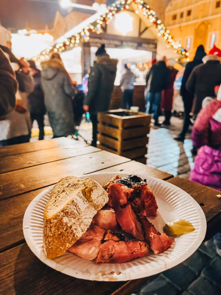 A close-up of a plate with sliced smoked meat, a crusty piece of bread, and a dollop of mustard, set on a wooden table at the Prague Christmas markets with festive lights and people in the background.
