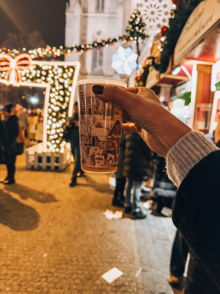 Woman holding a decorative holiday cup filled with hot mulled wine at Christmas markets in Prague, surrounded by glowing Christmas lights and festive decorations at night