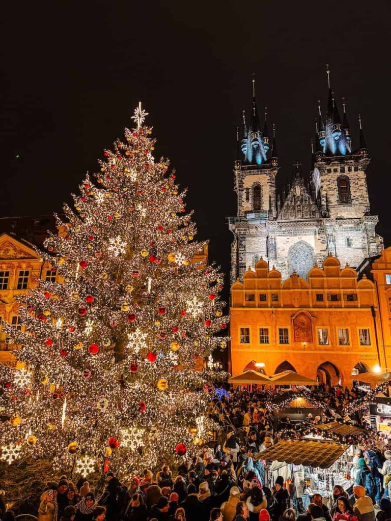 A massive Christmas tree covered in golden lights and red ornaments stands in Prague’s Old Town Square at night, surrounded by a crowd of people and wooden market stalls, with the illuminated Church of Our Lady before Týn in the background.