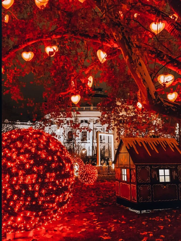 Enchanting night scene from Vienna’s Rathauspark with glowing red heart lights hanging from trees, oversized Christmas baubles, and a gingerbread-style hut illuminated in festive red.
