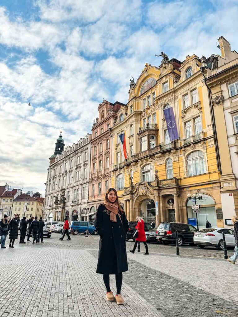 Woman standing in Prague’s Old Town Square with colorful Baroque and Rococo facades in the background on a bright winter day.