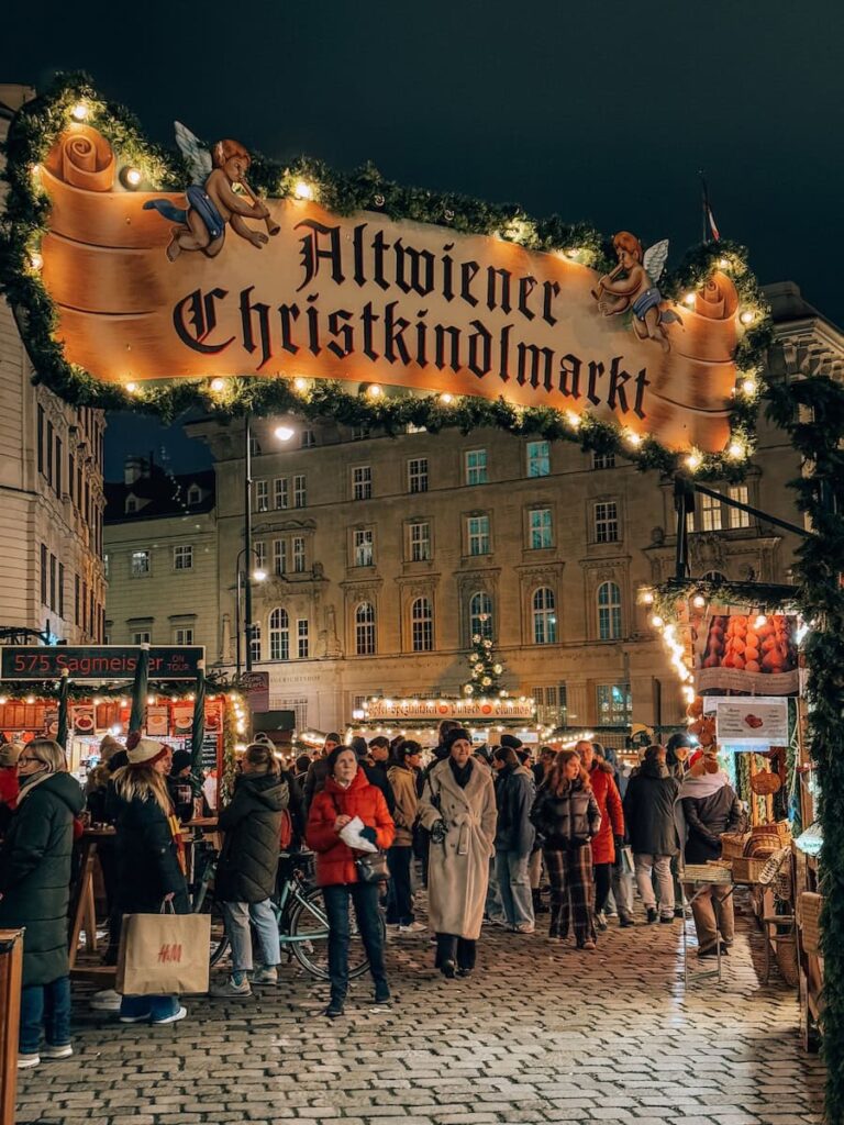 Crowds enjoying the festive atmosphere at the Altwiener Christkindlmarkt in Vienna, under a warmly lit entrance sign with garlands and angels.