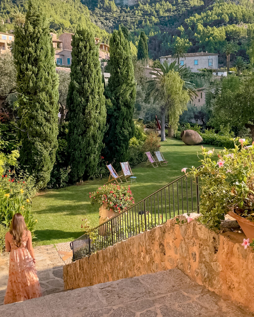 Woman in a flowing pink and cream maxi dress walking down stone steps into a lush garden with tall cypress trees, blooming flowers, and lounge chairs, surrounded by Mediterranean hillside homes in Deià, Mallorca.