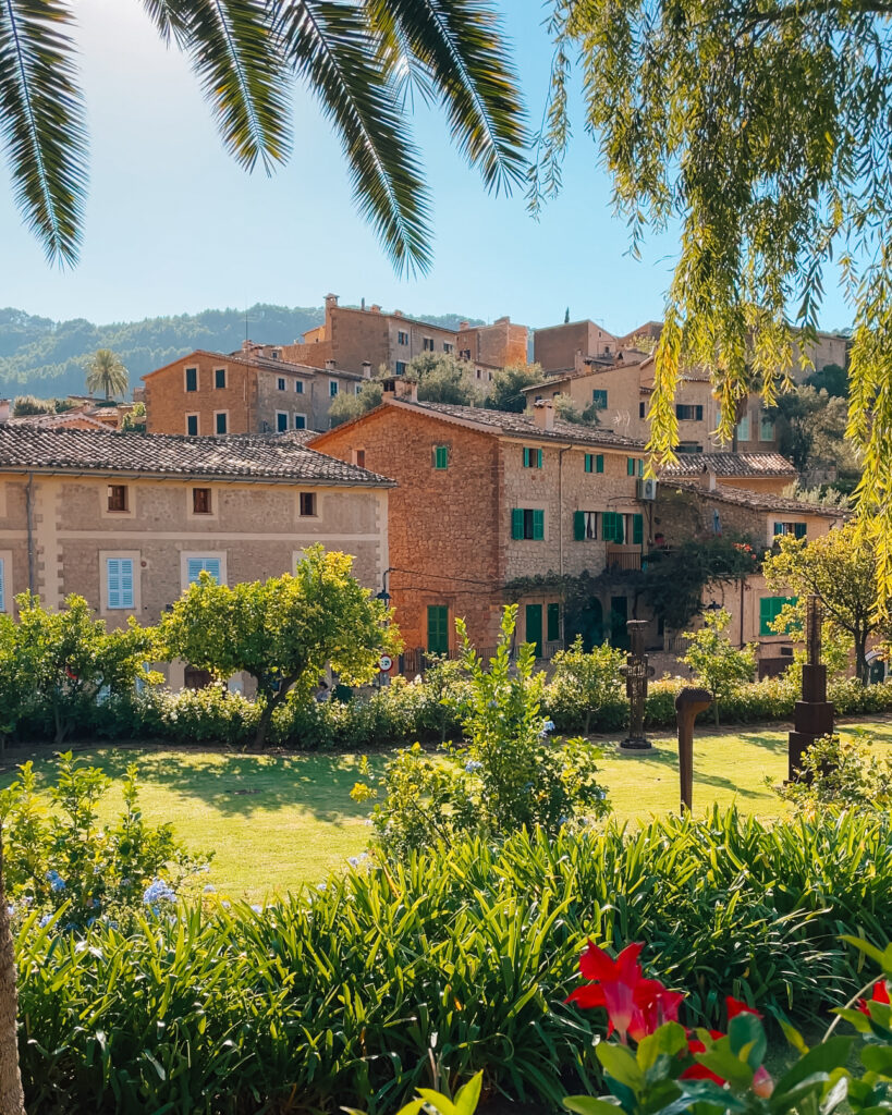 View of charming stone houses and lush gardens in Deià, Mallorca, framed by palm leaves with green hills in the background.