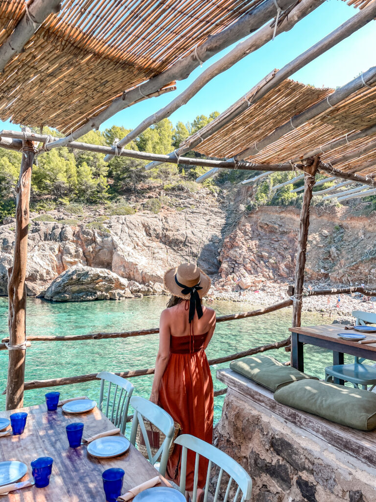 Woman in a rust-orange dress and straw hat standing on a rustic wooden terrace at a seaside restaurant in Cala Deià, Mallorca, overlooking turquoise waters and rocky cliffs.