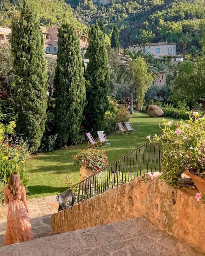 Woman in a flowing pink and cream maxi dress walking down stone steps into a lush garden with tall cypress trees, blooming flowers, and lounge chairs, surrounded by Mediterranean hillside homes in Deià, Mallorca.