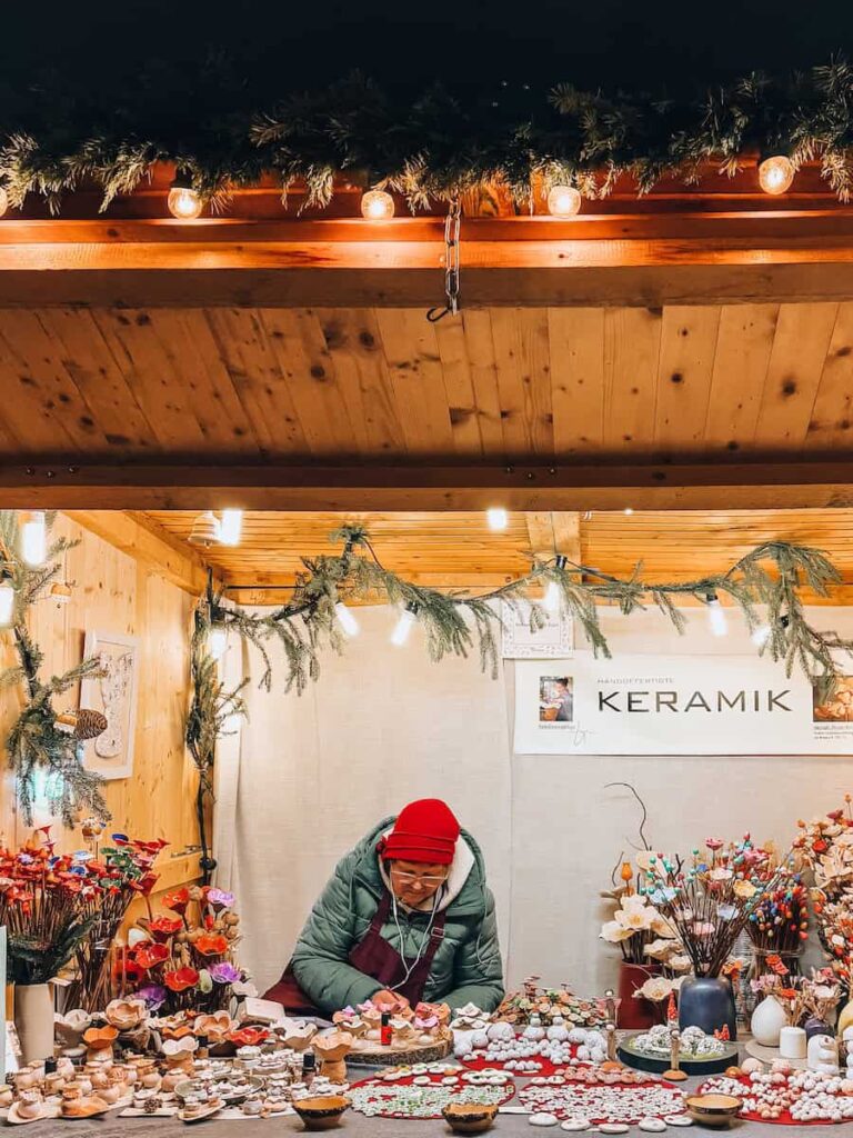 Elderly vendor selling handmade ceramic ornaments and floral decorations at a cozy Christmas market stall in Vienna, surrounded by twinkling lights and pine garlands.