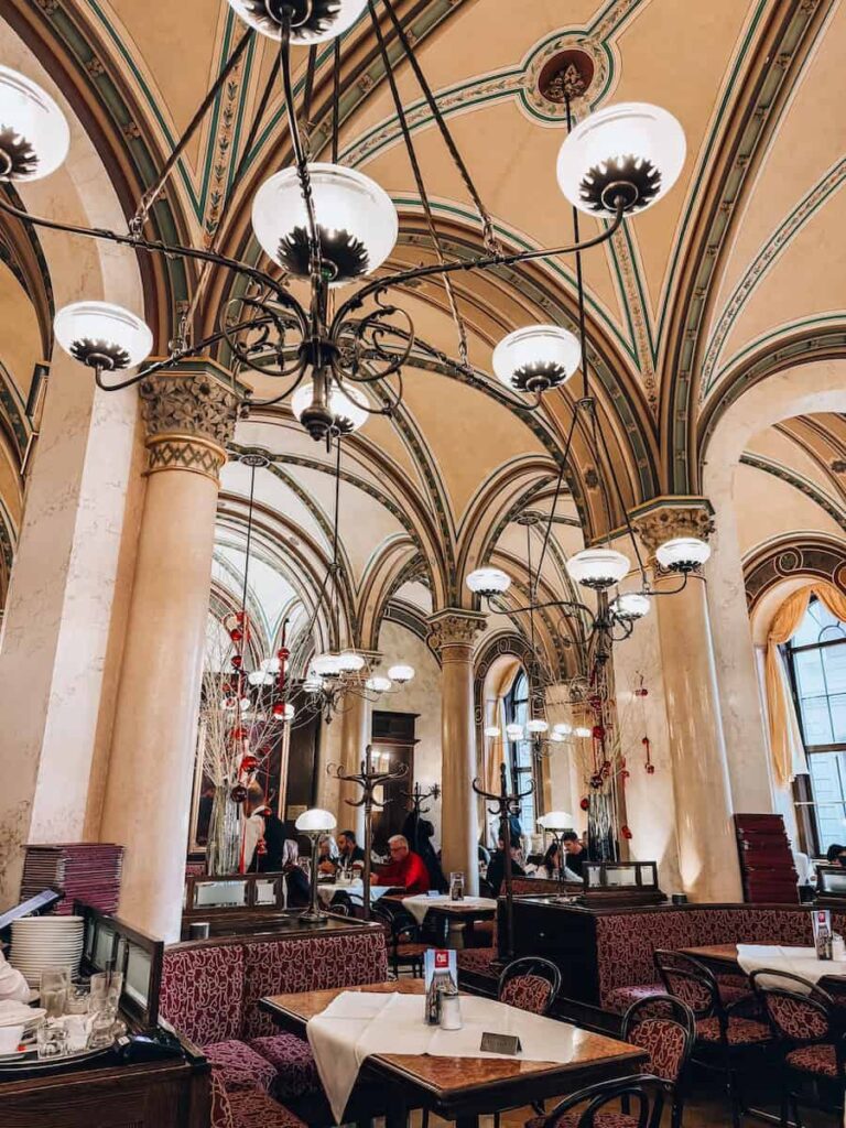 Elegant interior of Café Central in Vienna, showcasing vaulted ceilings with green and gold detailing, ornate hanging chandeliers, and cozy red-upholstered booths. Patrons sit at marble-top tables enjoying the refined ambiance of this historic Viennese coffeehouse.